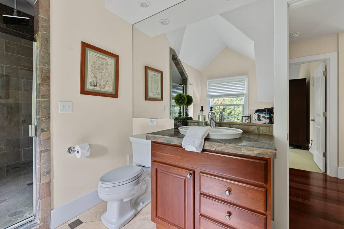 A bright bathroom with a dark wood vanity, a white vessel sink, and a large mirror extending to the ceiling. A toilet is next to the vanity, and a tiled walk-in shower with a glass door is visible on the left.