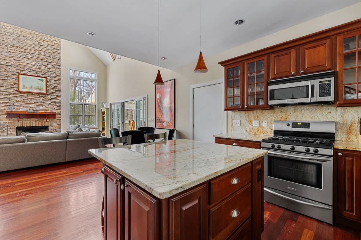 An open-concept view from the kitchen island shows the adjacent living room with a stone fireplace and a dining area with a table and chairs, all connected by rich hardwood flooring under vaulted ceilings.