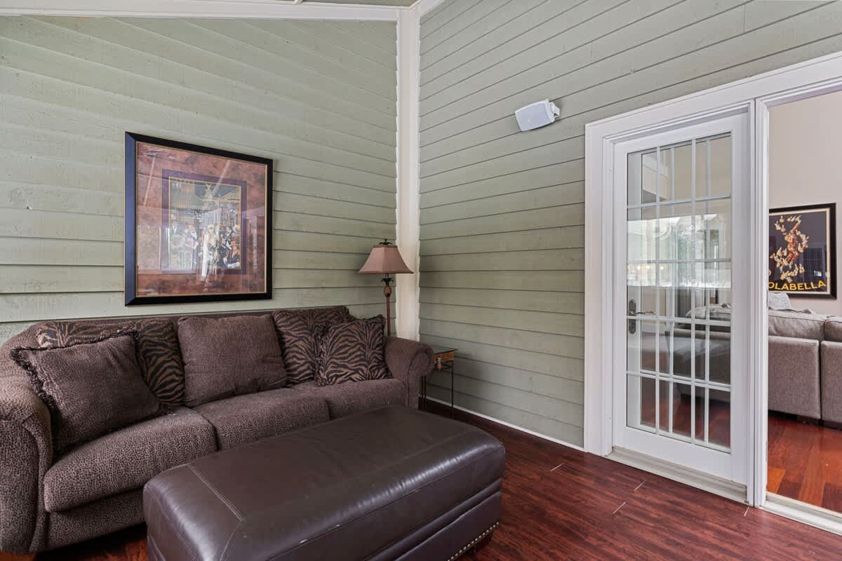 This cozy section of the enclosed porch features a comfortable brown sofa with an ottoman, set against light green shiplap walls, with a large framed artwork and a floor lamp, and a glass door leading to another room.