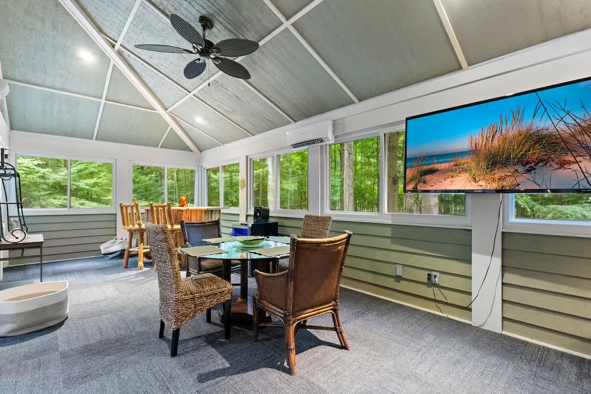 A bright screened-in porch features a vaulted ceiling with a fan, large windows overlooking trees, and two seating areas with a glass-top table and wicker chairs.