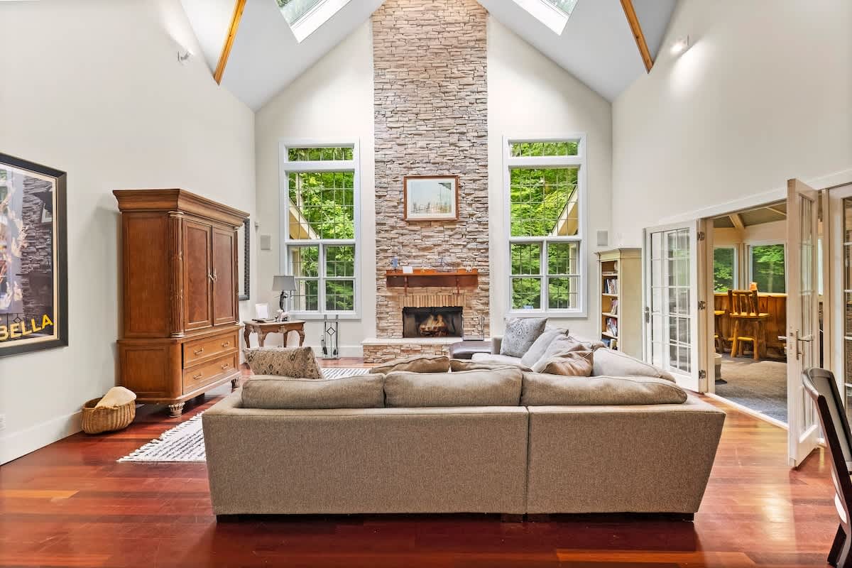 A spacious living room features a soaring vaulted ceiling with skylights, a floor-to-ceiling stone fireplace, and large windows overlooking lush greenery, complemented by a large sectional sofa and rich hardwood floors.