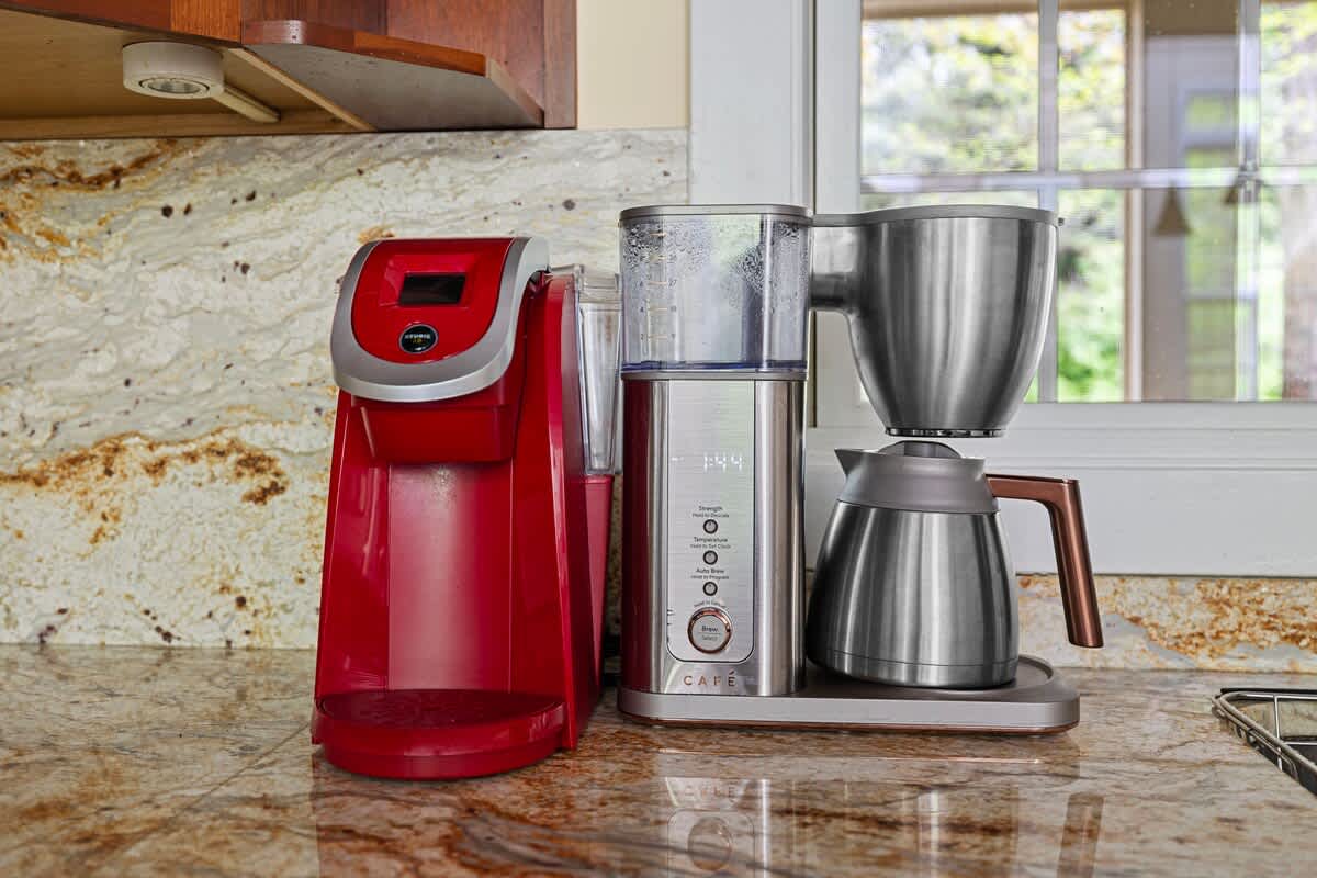A close-up of the kitchen counter displays a red single-serve coffee maker and a stainless steel drip coffee machine, resting on speckled granite countertops next to a window.