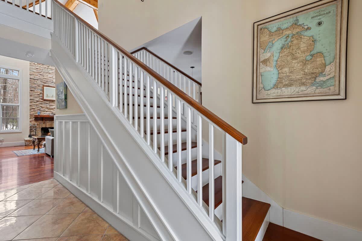 A grand staircase with white balusters and dark wood handrails, leading upstairs from a tiled floor area. A large framed map hangs on the light tan wall beside the stairs, adding a touch of classic decor.