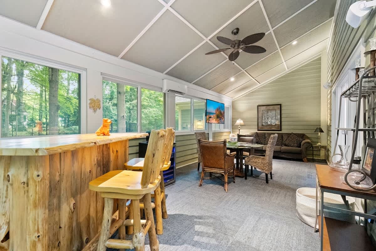 A bright enclosed porch features a rustic wooden bar with stools, a dining table, and a comfortable sofa, all surrounded by large windows offering views of the wooded outdoors, with a vaulted ceiling and ceiling fan.