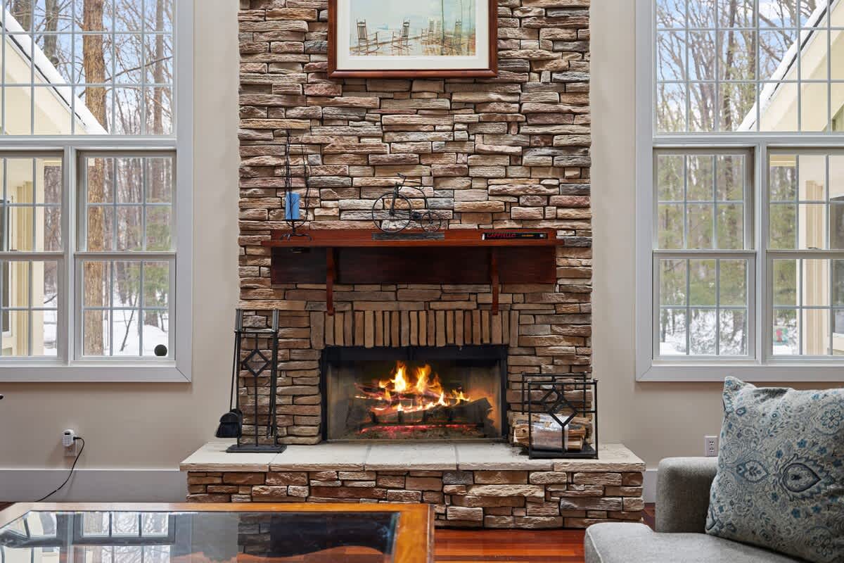 A cozy living room centered around a large stone fireplace with a roaring fire, flanked by two tall windows looking out to a snowy, wooded landscape. A comfortable sofa with patterned pillows is partially visible on the right.