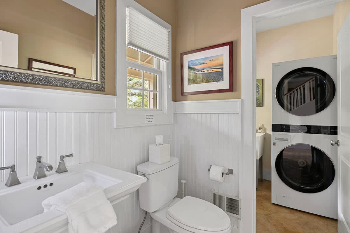 A compact bathroom featuring a white pedestal sink, a toilet, and wainscoting on the walls. A stacked washer and dryer unit is visible through an open doorway, indicating a combined laundry area for convenience.