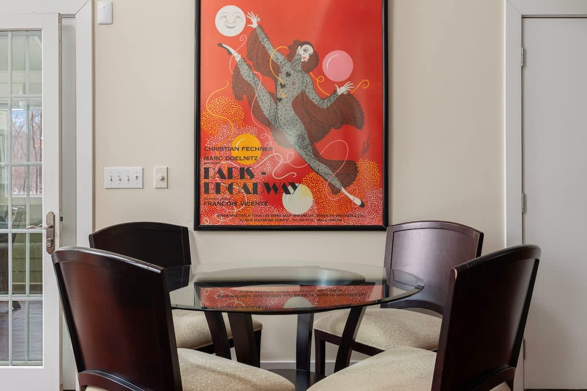 A cozy dining nook features a round glass-top table with four dark wood chairs, set against a light wall adorned with a vibrant red and orange art poster.