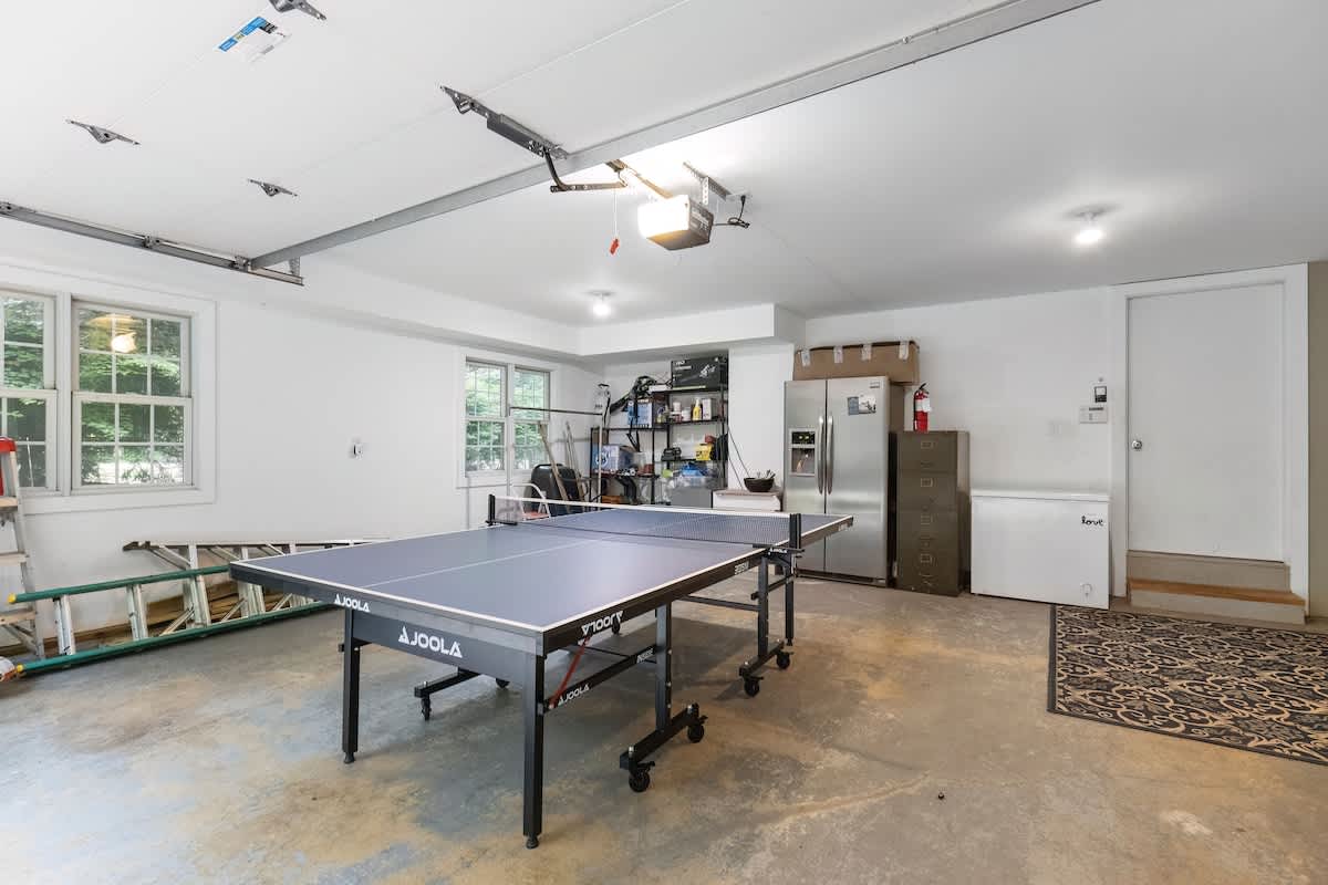 A spacious garage converted into a recreation area, featuring a blue JOOLA ping pong table in the center. Storage shelves, a stainless steel refrigerator, and a freezer are visible along the back wall, with windows providing natural light.