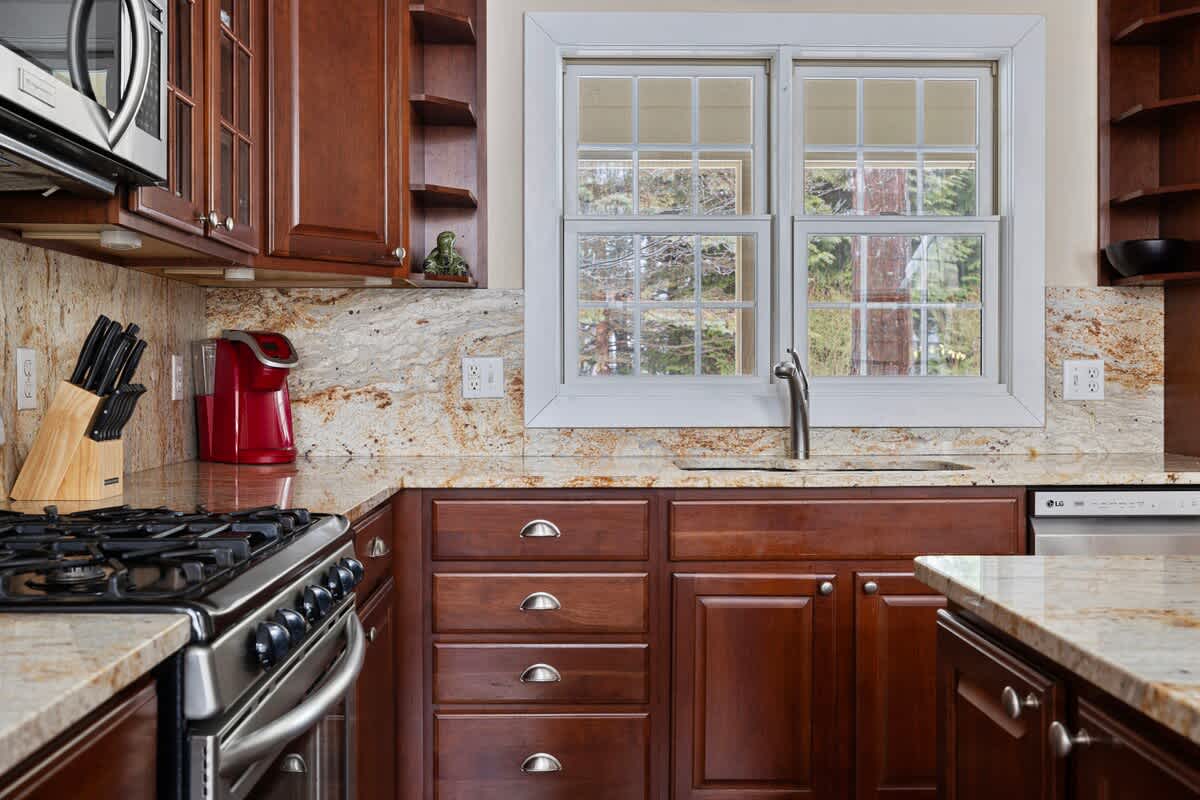 A detailed view of the kitchen counter reveals granite countertops, a stainless steel sink beneath a large window, and a gas range with a microwave above, surrounded by dark wood cabinetry.