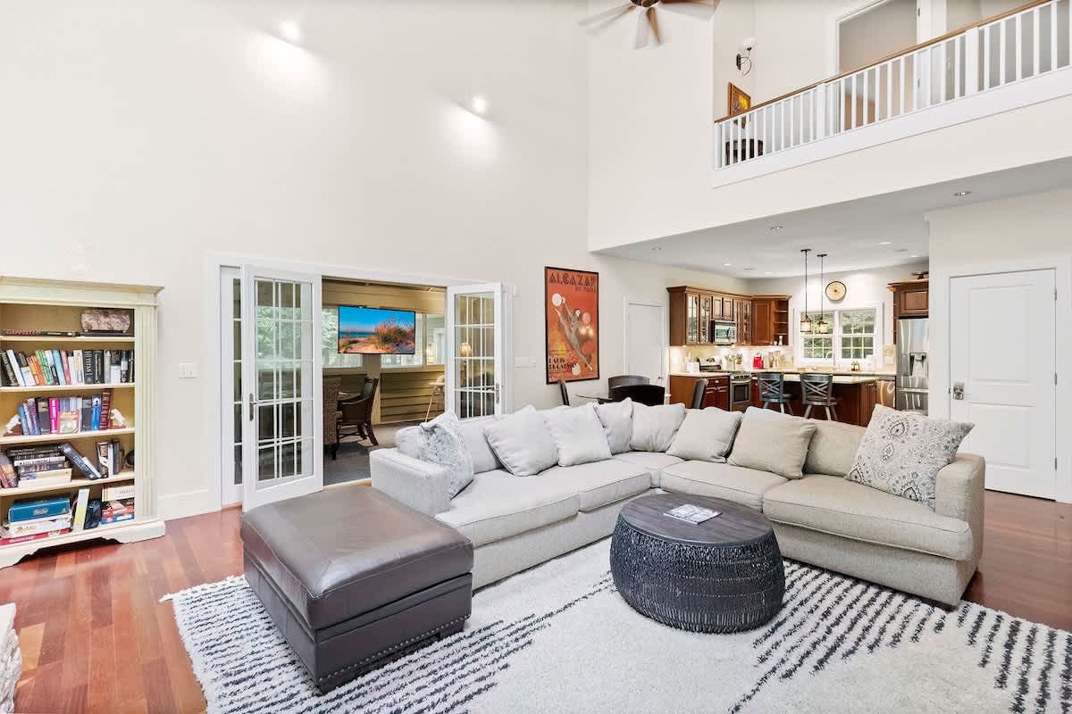 An open-concept living area features a large sectional sofa and ottoman on a patterned rug, with a view into the kitchen and a screened porch through French doors.