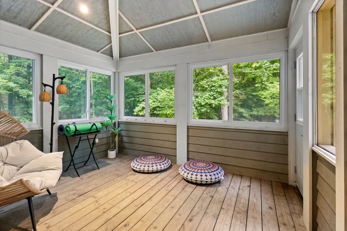 A serene corner of the enclosed porch is set up for relaxation or exercise, featuring a yoga mat on a stand, floor cushions, and a wicker chair, with large windows providing ample natural light and views of the surrounding trees.