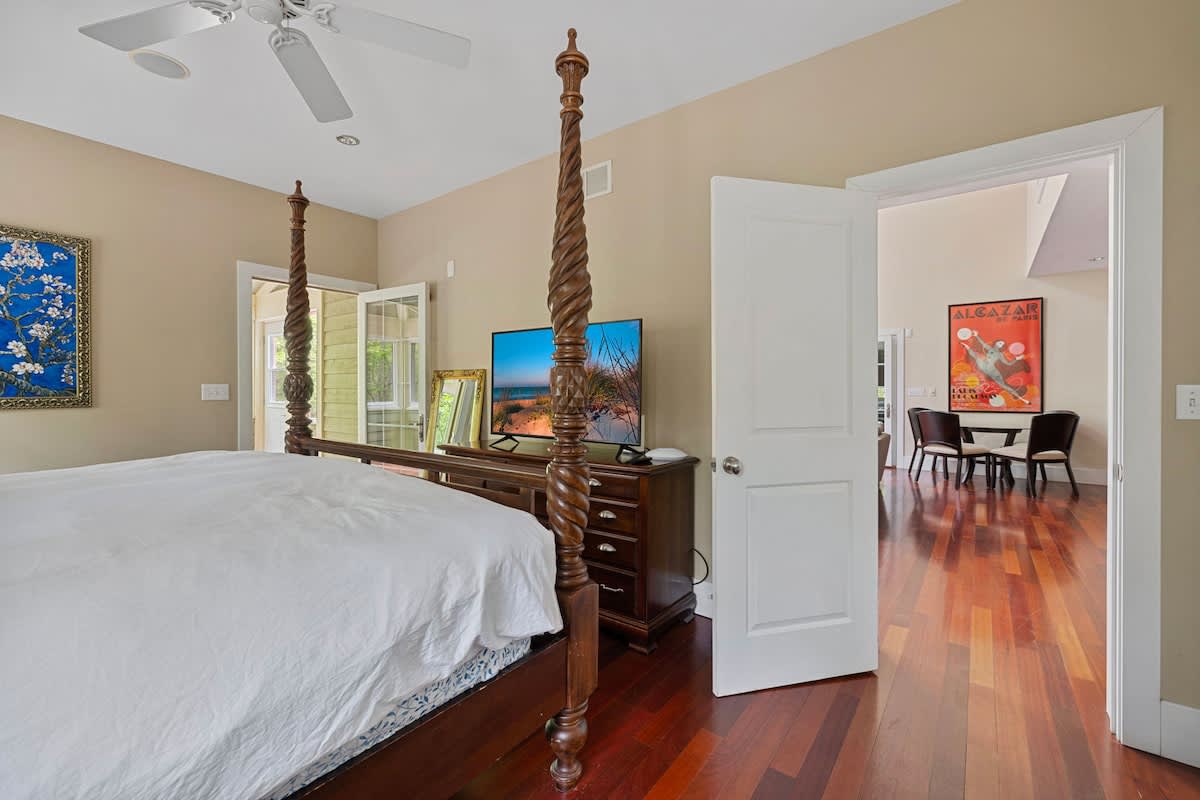 View from the foot of a dark wood four-poster bed in a bedroom with hardwood floors, showing a dresser with two flat-screen TVs. French doors lead to an outdoor space, and a doorway opens to another interior room.