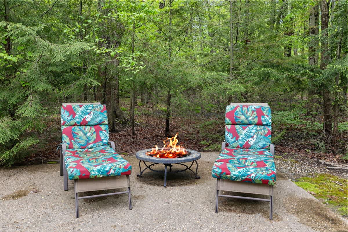 Two comfortable lounge chairs with vibrant tropical print cushions are arranged around a lit fire pit on a concrete patio, nestled within a dense, lush green forest.