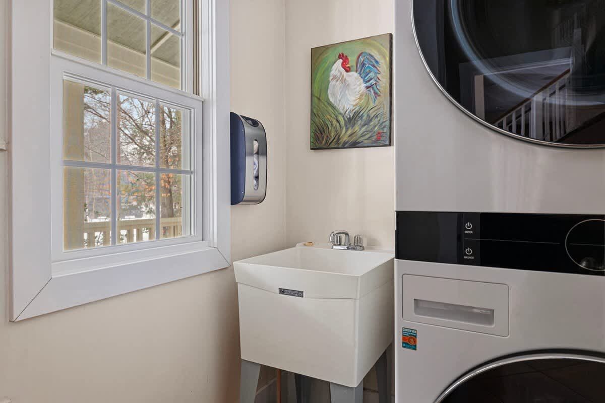 A bright laundry area featuring a white utility sink, a stacked washer and dryer unit, and a window with white grilles looking out to a snowy landscape. A colorful rooster painting hangs on the wall.