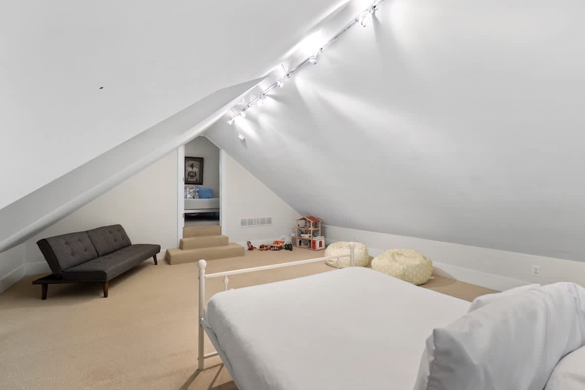 A spacious attic bedroom with white sloped ceilings and light carpet, featuring a white metal bed frame and a grey futon-style sofa. An arched opening leads to another room, and track lighting illuminates the space.