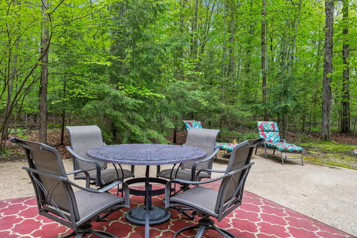 A round outdoor dining table with four chairs sits on a red patterned rug, with additional lounge chairs and a fire pit visible in the distance, all nestled within a lush green forest.