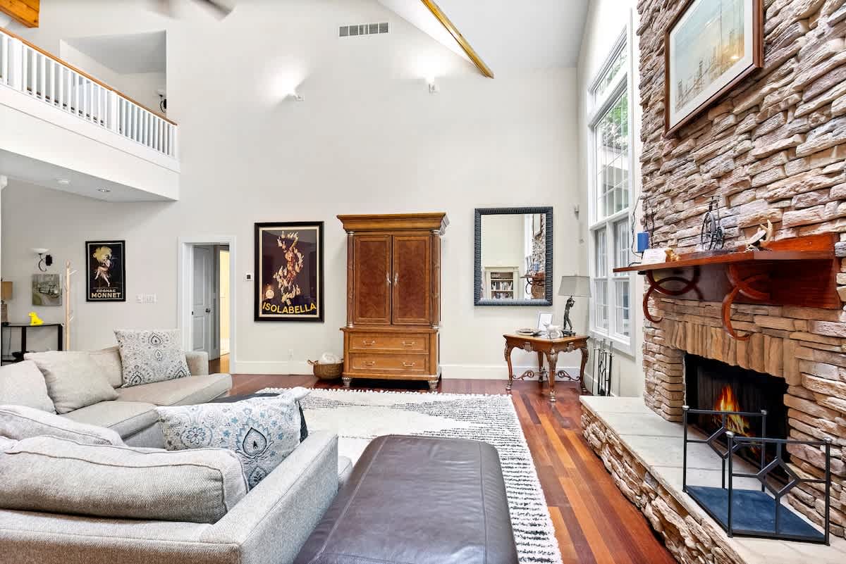 This expansive living room boasts a vaulted ceiling, a prominent stone fireplace with a wooden mantel, and dark hardwood floors, with a large sectional sofa and a tall wooden armoire, and a glimpse of a loft area above.