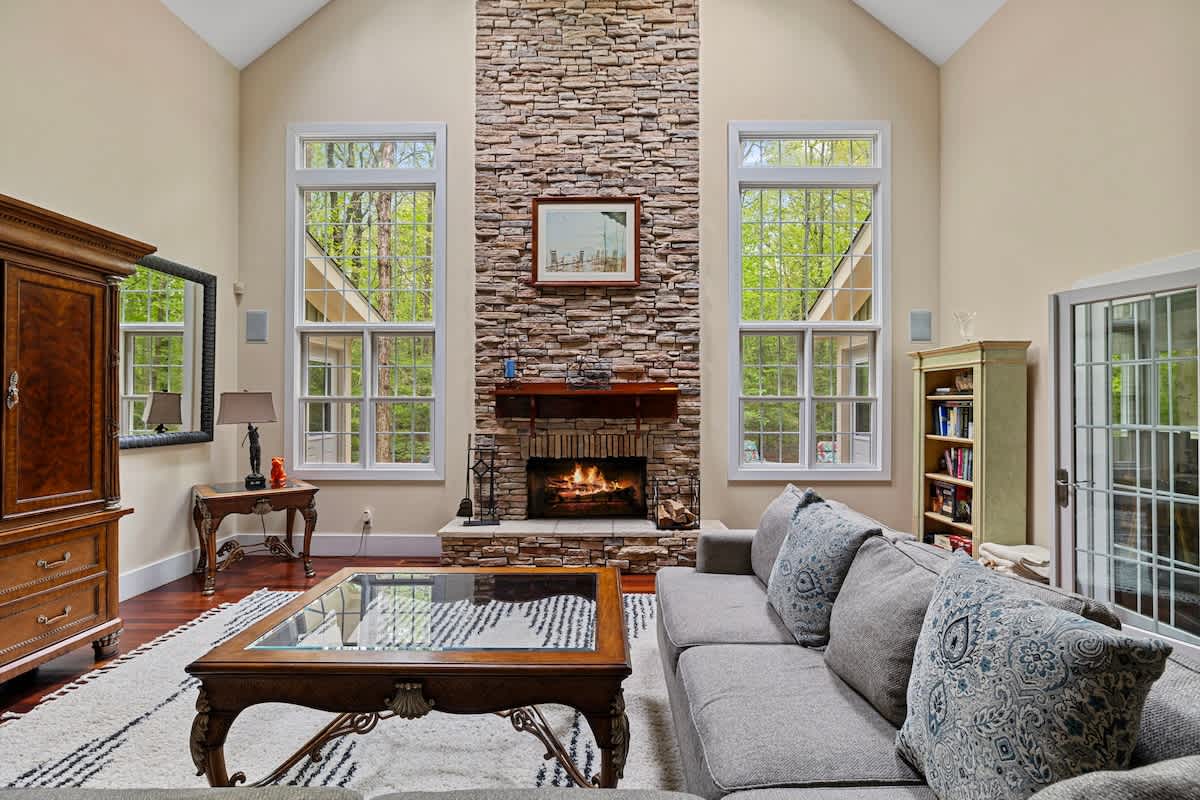 A spacious living room boasts a tall stone fireplace with a mantel, flanked by two large windows offering views of lush trees, and a comfortable gray sofa on a white shag rug.