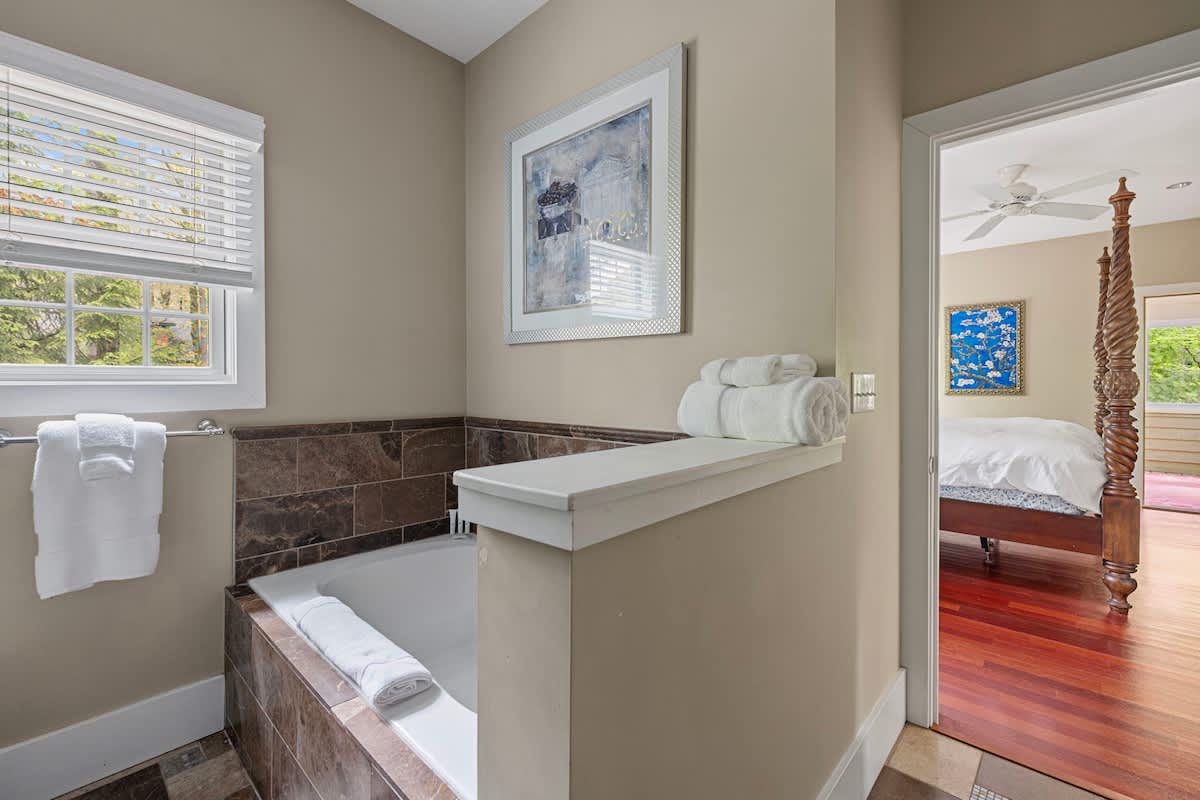 A section of a bathroom featuring a built-in bathtub with dark tiled surround, a window with blinds, and light tan walls. Fresh white towels are neatly stacked on a shelf and by the tub, suggesting a relaxing soak.