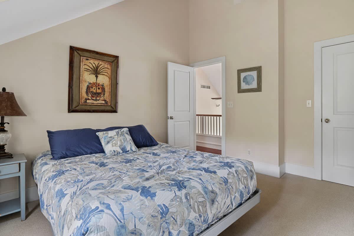 A bedroom with a high vaulted ceiling and light beige walls, featuring a bed with a blue and white floral duvet and dark blue pillows. A framed palm tree artwork hangs above the bed, and a small light blue side table with a lamp is visible.