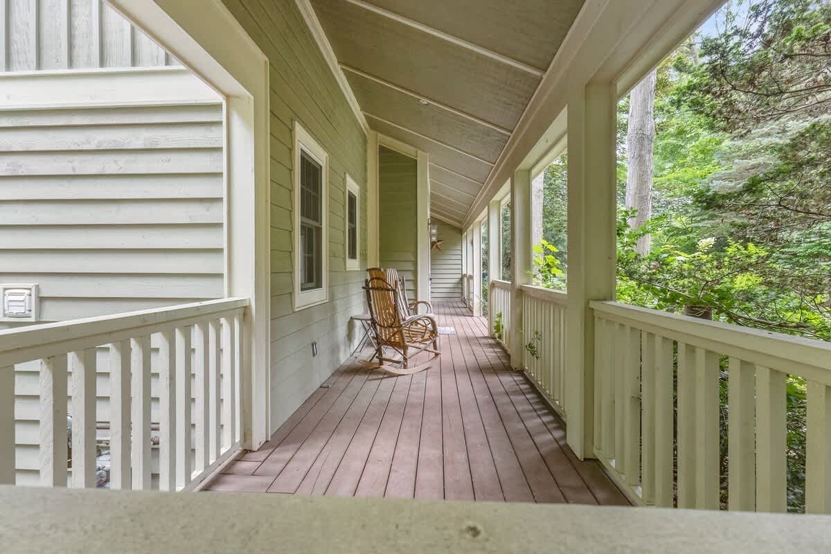 A long, covered porch with light green siding and white railings, featuring two wooden rocking chairs. The porch overlooks a lush, wooded area, providing a serene outdoor seating space.