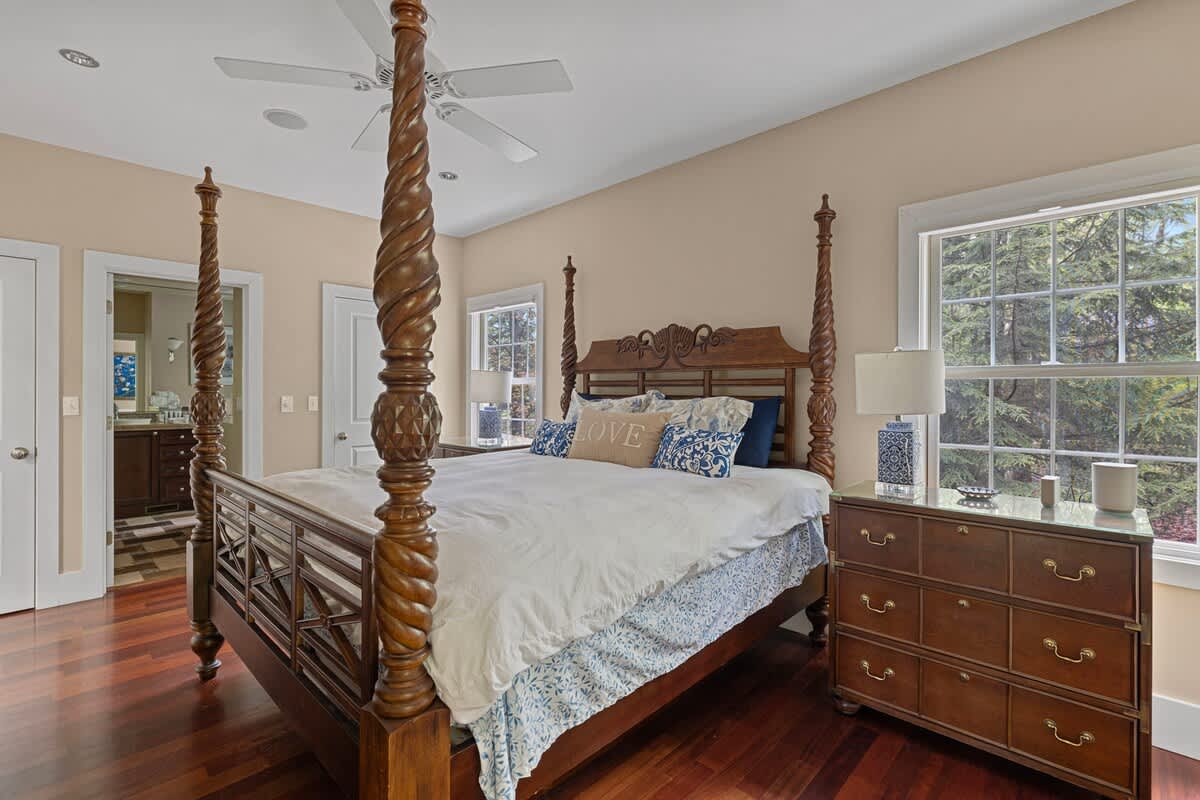 A wide shot of a bedroom with a dark wood four-poster bed, white bedding, and blue patterned pillows, complemented by dark hardwood floors and light tan walls. A dresser with a lamp sits by a large window, and a doorway leads to another room.
