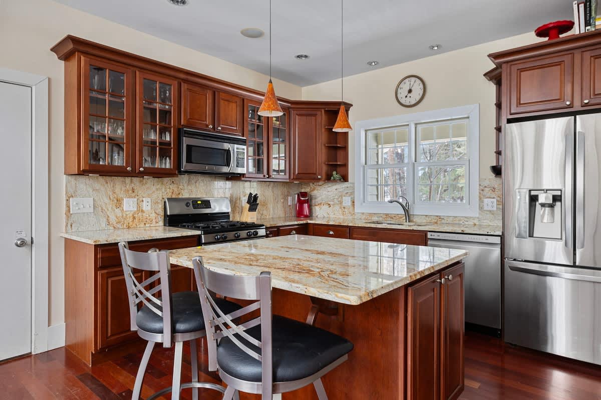 A modern kitchen features rich cherry wood cabinetry, granite countertops, stainless steel appliances, and a central island with two bar stools, illuminated by pendant lights.