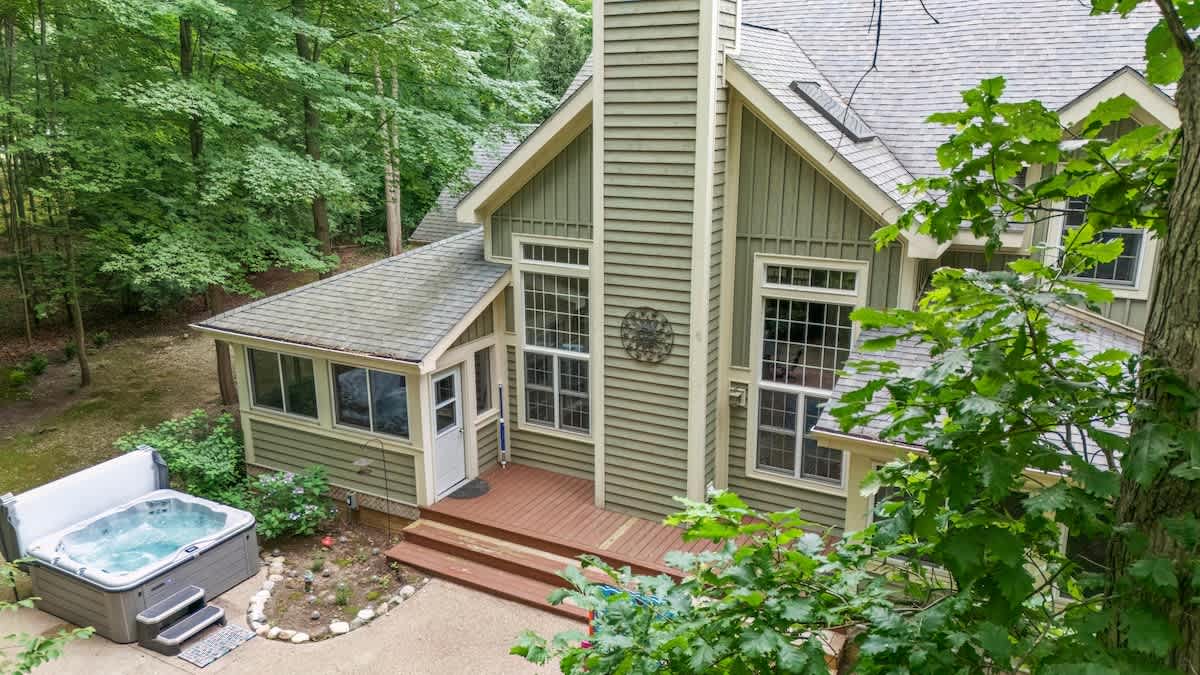 An elevated view of the green-sided house's rear, showcasing a hot tub on a concrete patio adjacent to a wooden deck, with a tall chimney and numerous windows, all enveloped by a vibrant green forest.