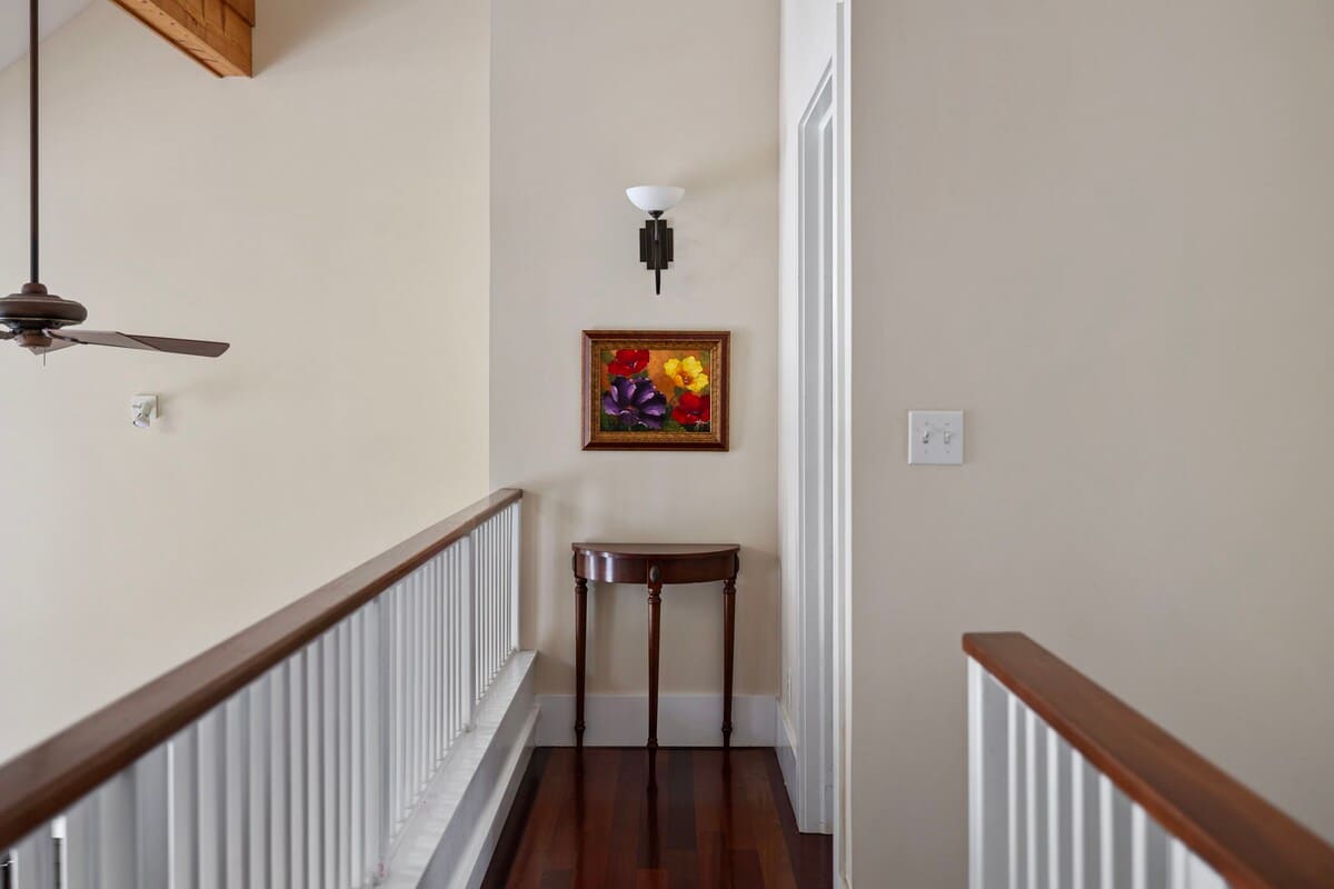 An upstairs hallway with light tan walls, dark hardwood floors, and a white railing overlooking the space below. A small console table with a vibrant floral painting above it adds a decorative touch.