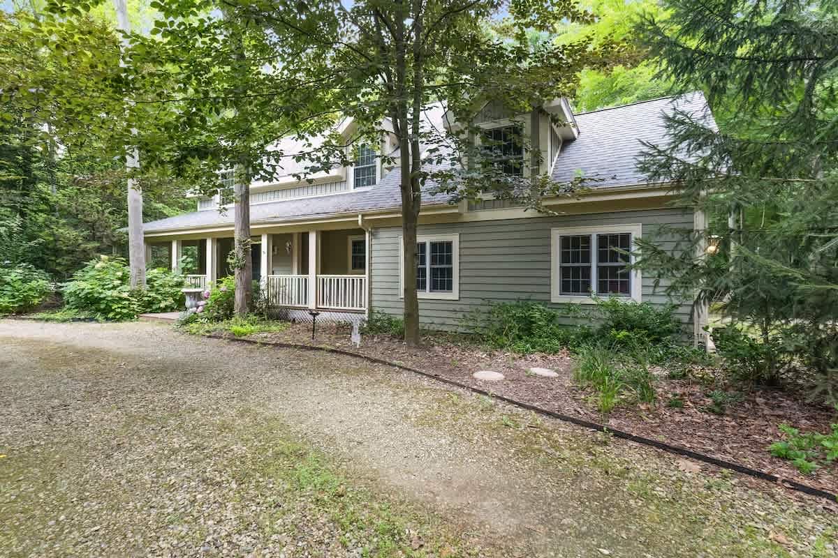 The front of the green-sided house features a gravel driveway leading to a welcoming covered porch with white railings, nestled among mature green trees.
