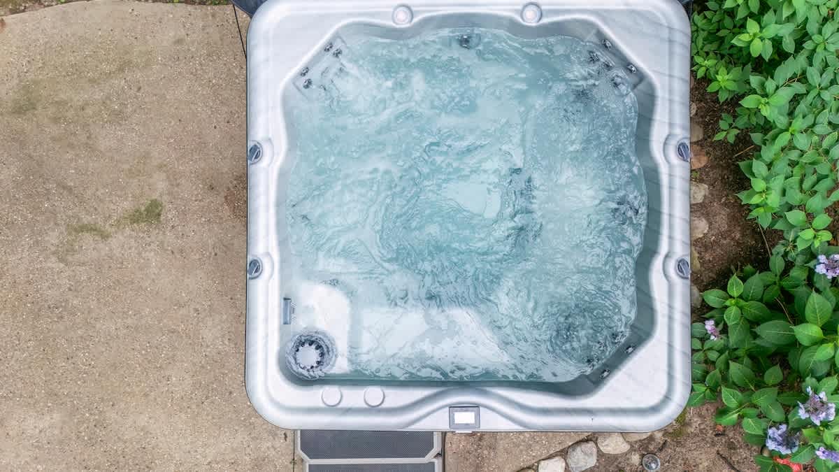 An overhead view captures a bubbling gray hot tub set on a concrete patio, surrounded by vibrant green foliage and small purple flowers.