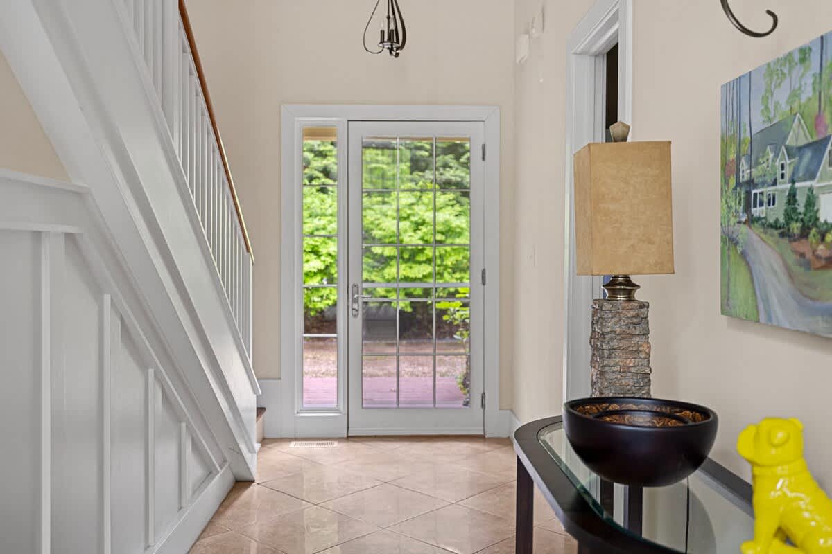 A welcoming foyer features light tiled flooring, a white staircase with wainscoting, and a glass-paned front door offering a view of outdoor greenery.