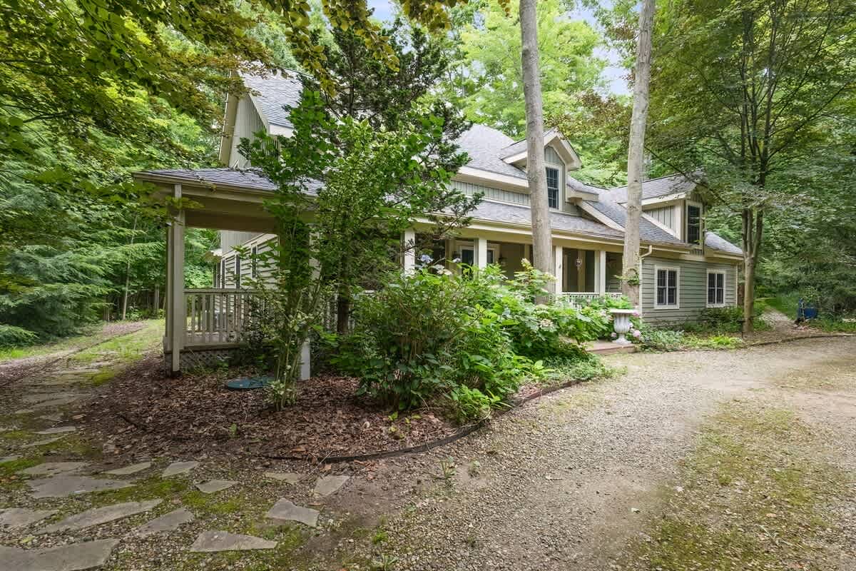 The front exterior of a light green house with multiple gables and dormer windows is nestled among lush trees, featuring a covered porch and a gravel driveway.