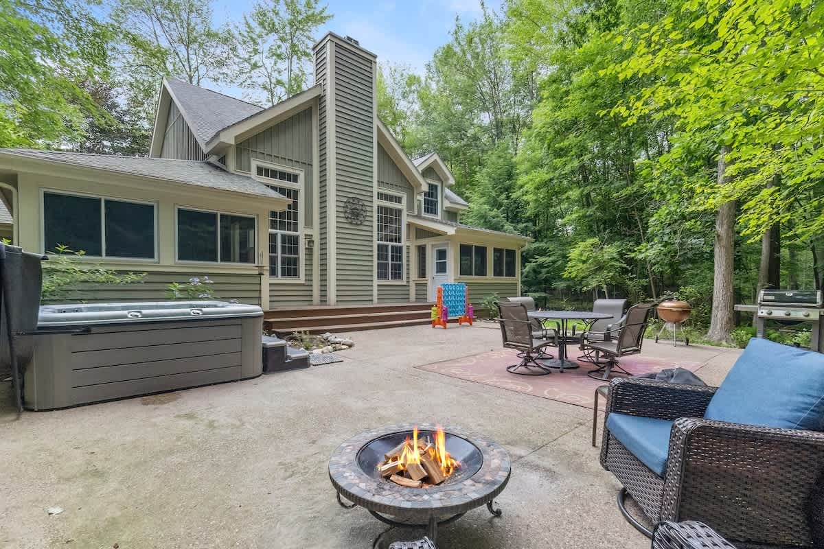 An expansive concrete patio features a hot tub, a lit fire pit, an outdoor dining set on a red patterned rug, and a BBQ grill, all set against the green-sided house and surrounded by dense woods.