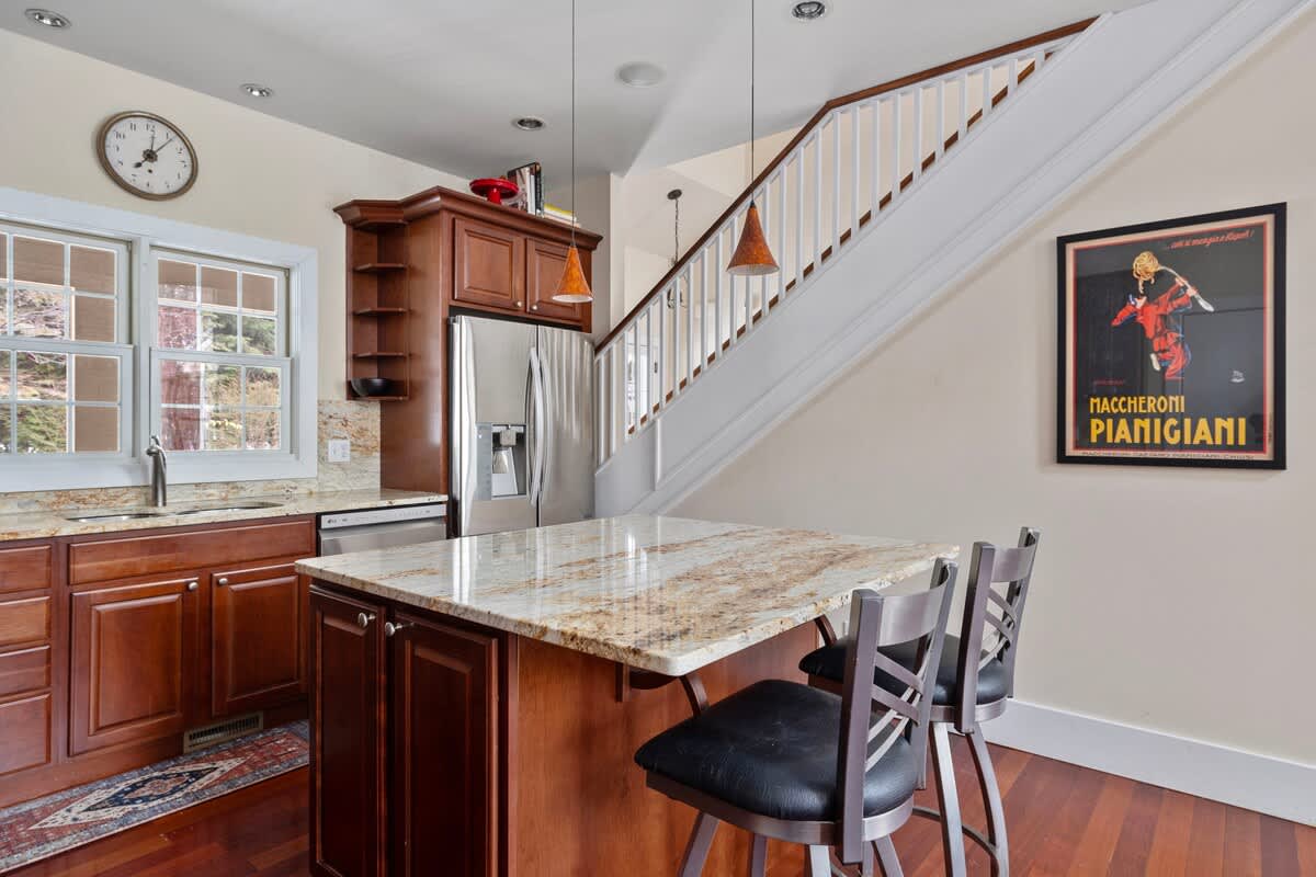 The kitchen showcases dark wood cabinets and a granite-topped island with seating, alongside a stainless steel refrigerator and a staircase leading upstairs, all illuminated by pendant lights and natural light from a window.