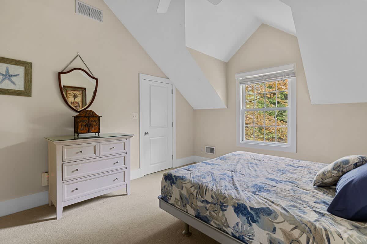 A cozy bedroom with a sloped ceiling, light beige walls, and a bed with a blue and white floral duvet. A light-colored dresser with a decorative mirror above it sits against one wall, next to a window overlooking autumn trees.