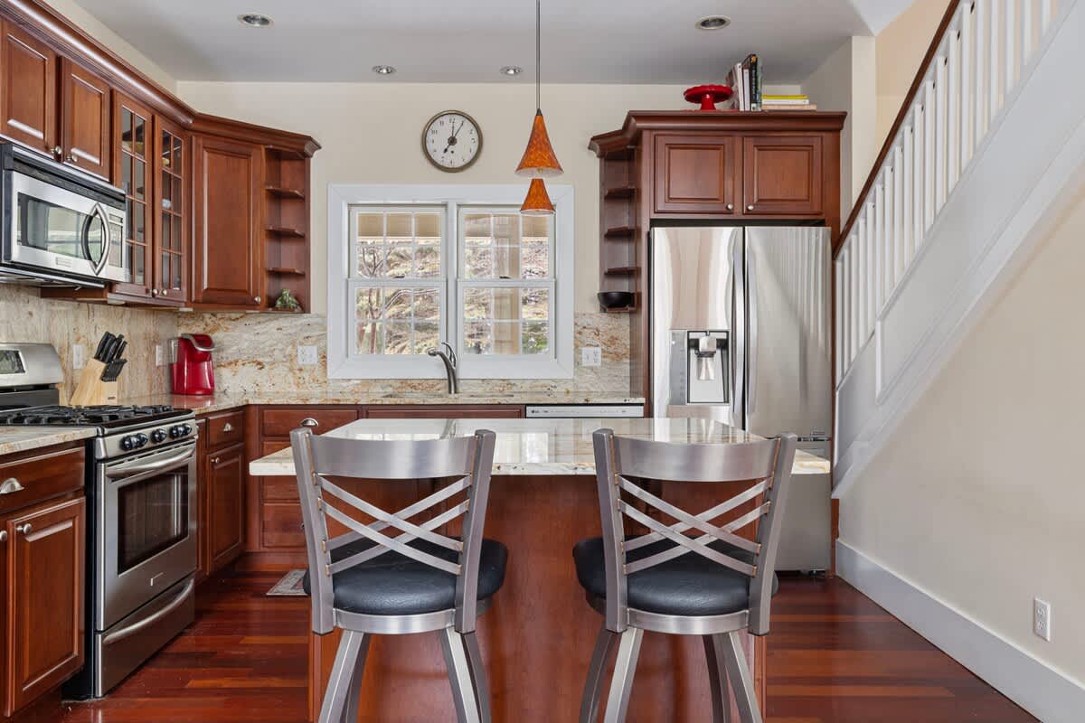 A well-appointed kitchen features dark cherry wood cabinetry, granite countertops, stainless steel appliances including a range and refrigerator, and a central island with bar seating, all set on polished hardwood floors.