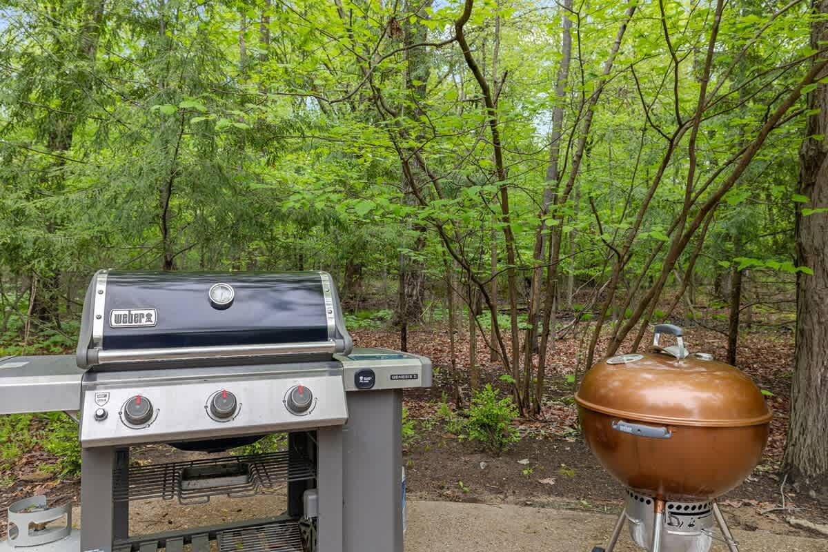 A modern black gas grill and a classic copper charcoal grill are positioned side-by-side on a concrete patio, ready for outdoor cooking amidst the surrounding green trees.