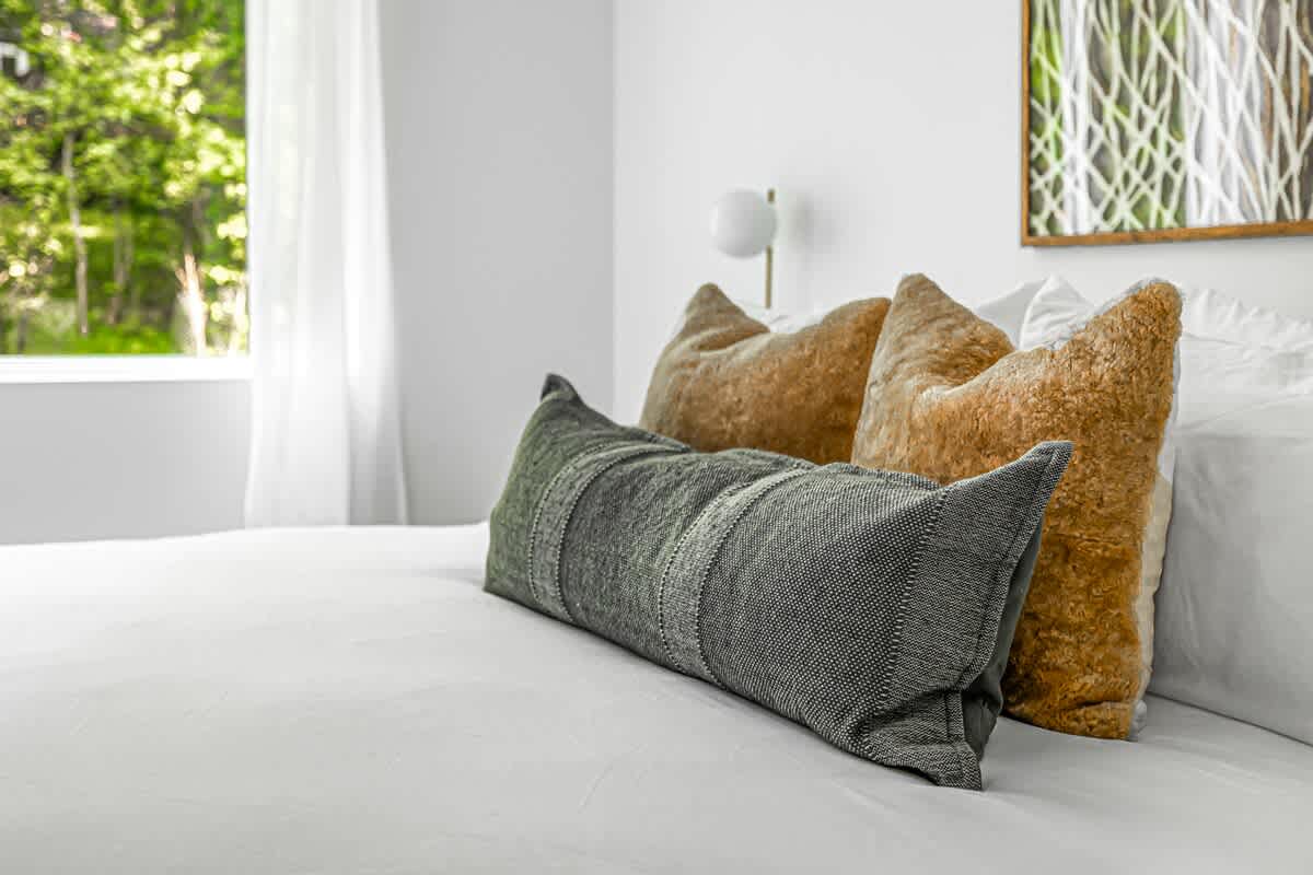 Close-up of a bed with crisp white bedding, featuring a long grey textured pillow and two fluffy brown accent pillows. A window with sheer curtains is visible in the background.