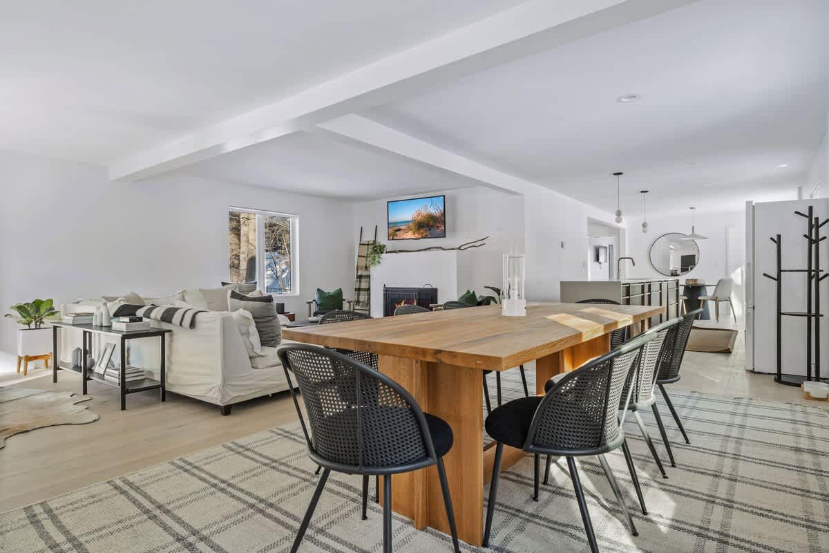 Expansive open-concept living space with light wood floors, white walls, a cozy fireplace, and a large wooden dining table with black chairs, leading into a modern kitchen.