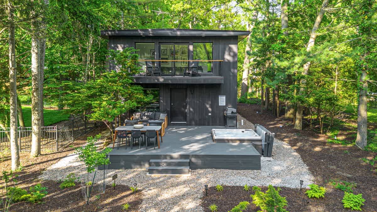 Modern black two-story house nestled among lush green trees, featuring a spacious back deck with an outdoor dining table, a hot tub, and a BBQ grill, all set on a gravel and landscaped yard.