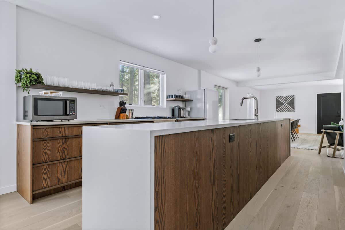 Contemporary kitchen showcasing a long island with a white countertop and dark wood paneling, a stainless steel sink, and a microwave on the counter with open shelving above.