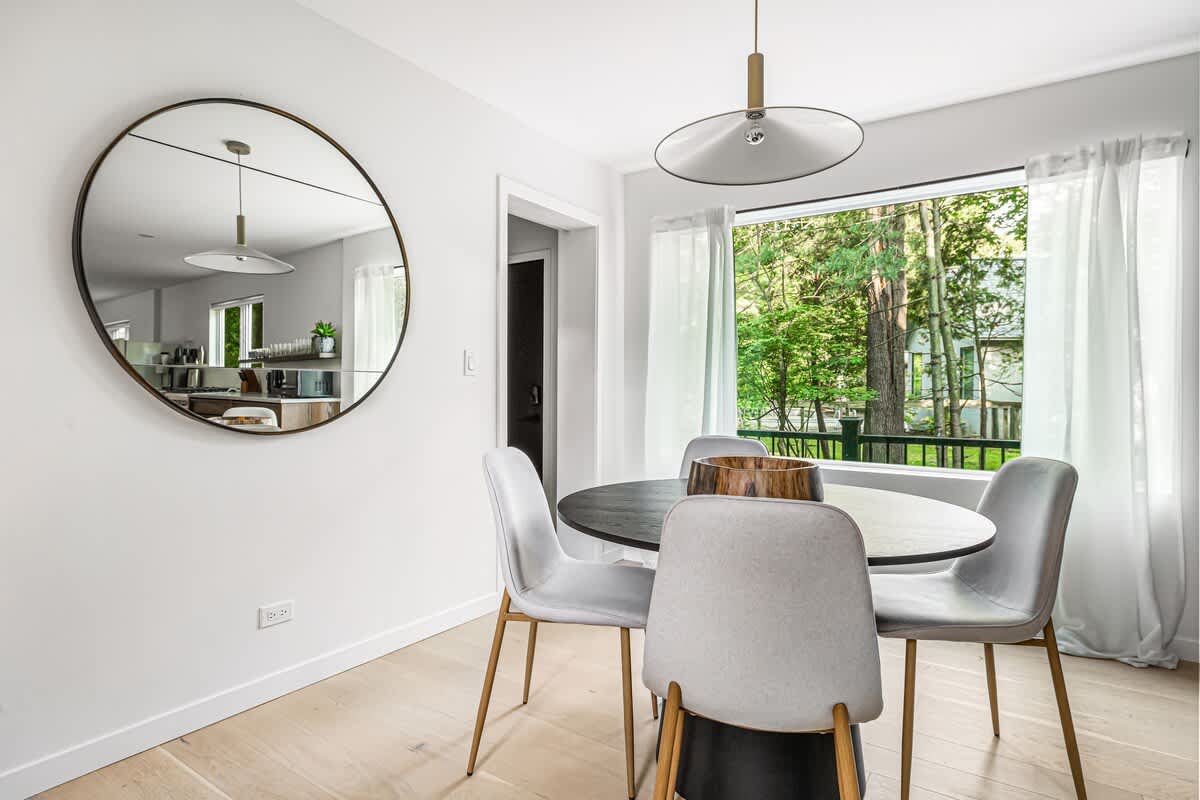 Cozy breakfast nook featuring a round black table, light grey chairs, a large circular mirror reflecting the adjacent kitchen, and a window with white curtains.