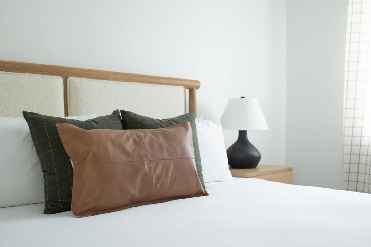 Close-up of a bed with a wooden and upholstered headboard, crisp white bedding, and a mix of green and brown accent pillows. A black table lamp sits on a wooden nightstand beside the bed.