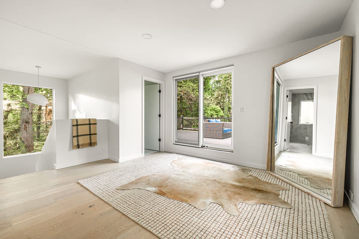 Bright, minimalist landing area with light hardwood floors, a large patterned rug, and a cowhide accent rug. A full-length mirror leans against the wall, and a sliding glass door opens to an outdoor deck surrounded by trees.