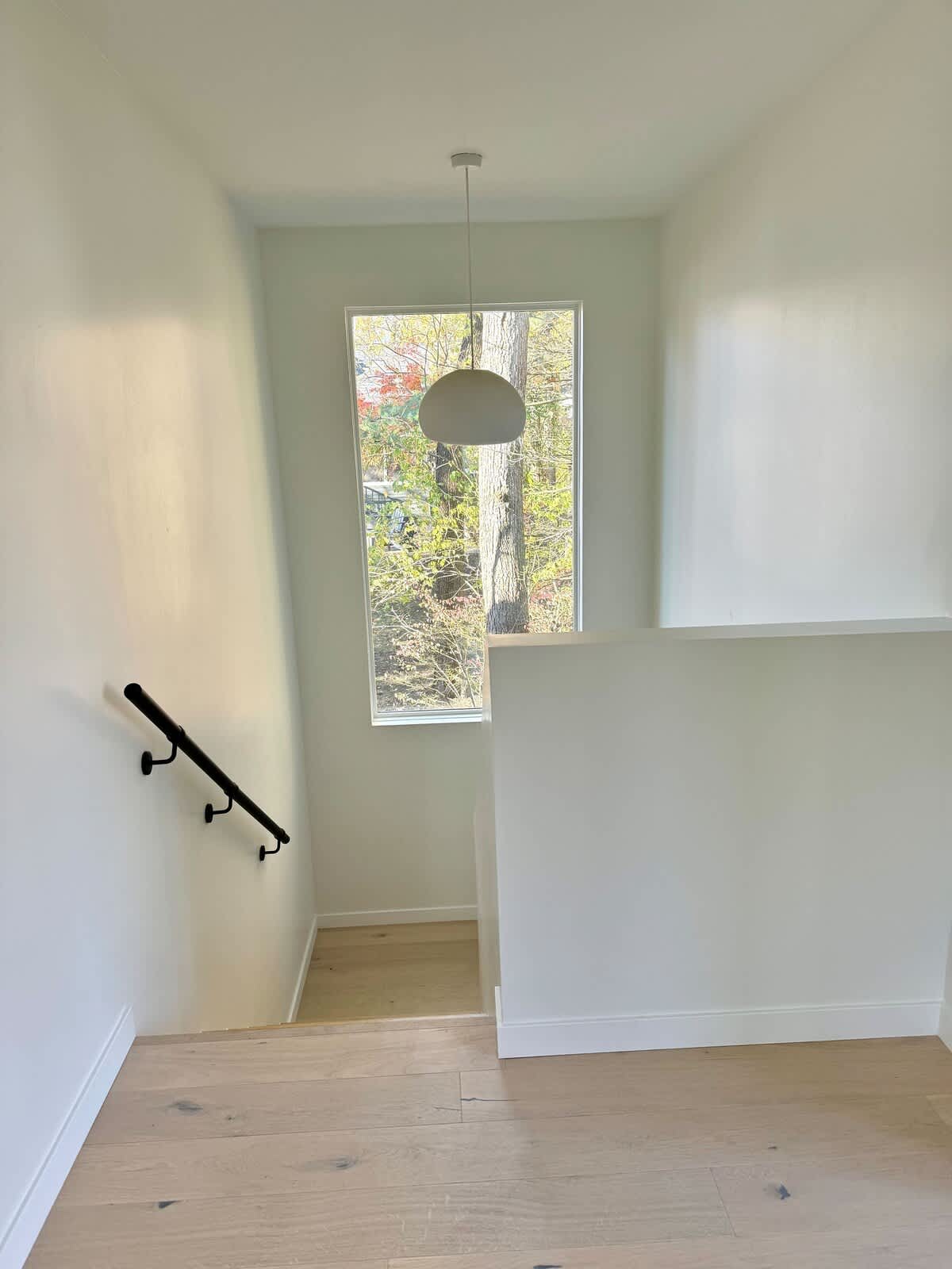 A bright, minimalist staircase with light wood steps and a black handrail, leading down from a landing. A large vertical window at the landing provides natural light and a view of outdoor trees.
