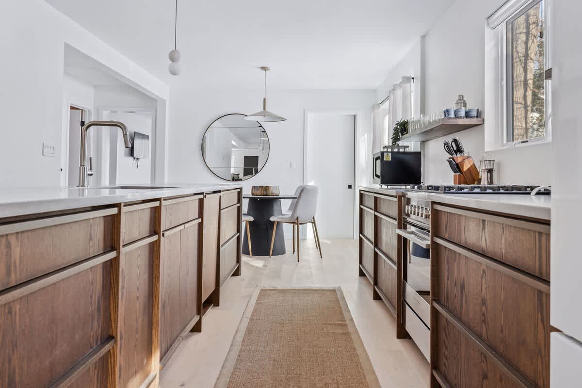 View down a galley kitchen with dark wood cabinetry and white countertops, leading to a bright breakfast nook with a round table and a large circular mirror on the wall.