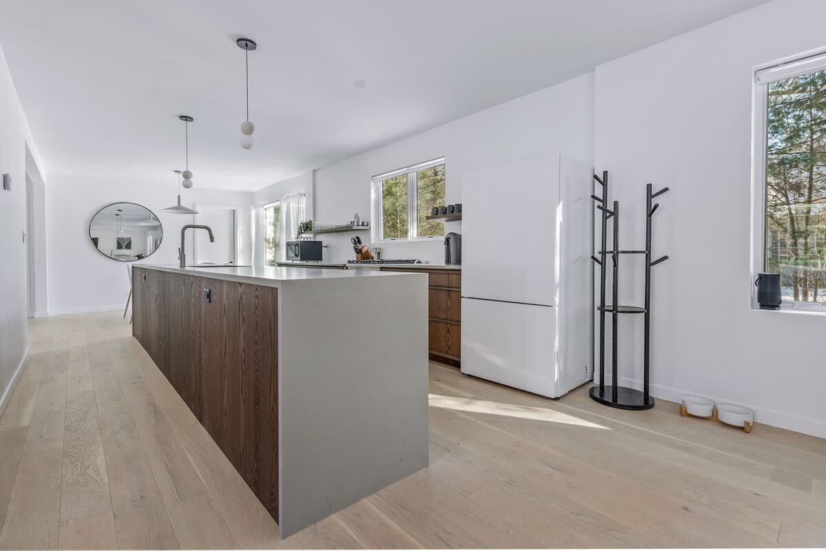 Spacious kitchen with a long island featuring a white countertop and dark wood paneling, light wood floors, a white refrigerator, and a modern black coat rack.