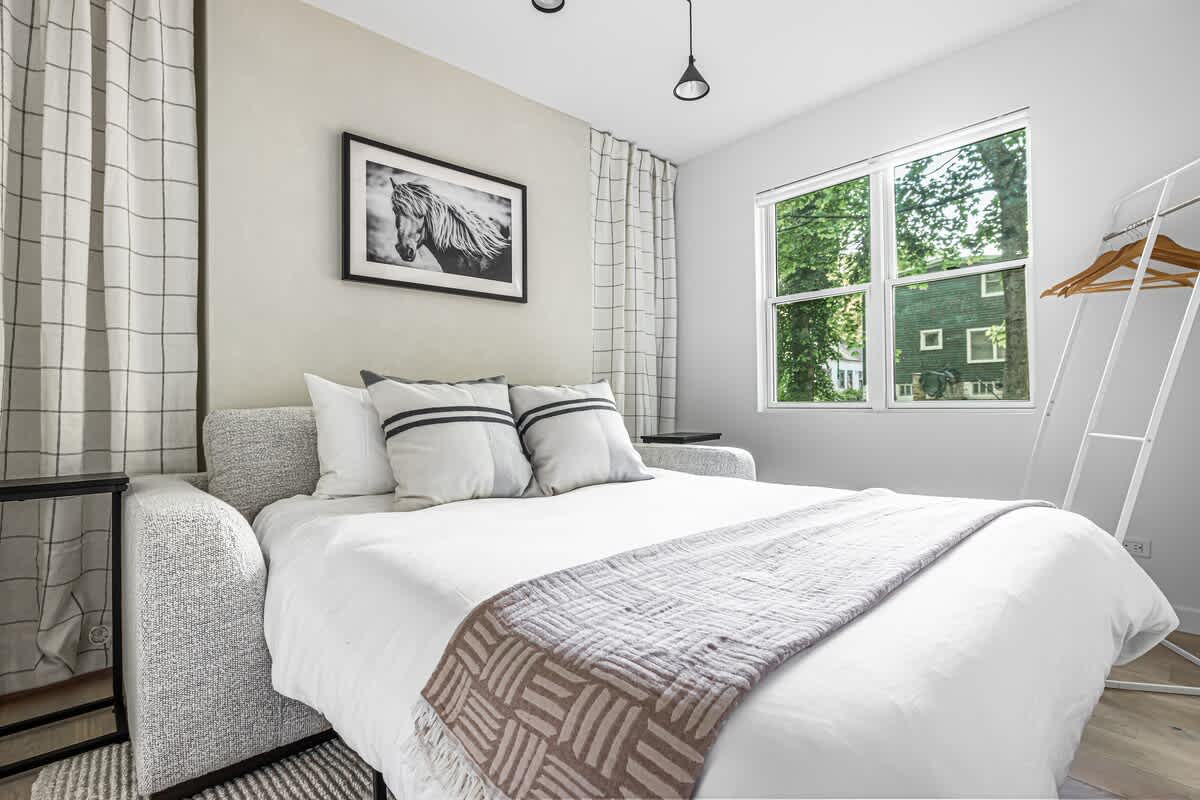 The light gray sofa from the previous image is now converted into a bed, dressed with white linens and a patterned gray throw, creating a cozy guest bedroom. A large window provides natural light and a view of trees.