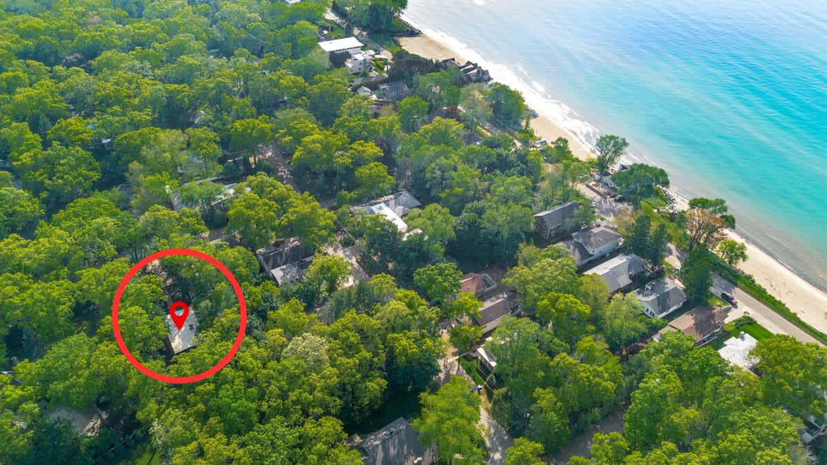 A high aerial perspective shows the property, marked by a red pin, situated within a lush green residential area, with the vibrant blue water and sandy shoreline of a large lake or ocean visible in the distance.